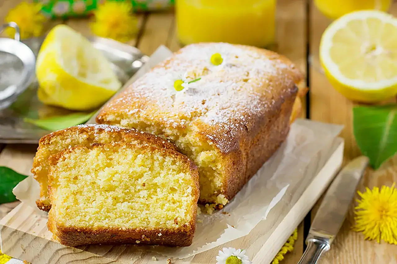 Un soffice plumcake al limone, spolverato di zucchero a velo, pronto per essere gustato. Accanto, spicchi di limone fresco e fiori gialli.
