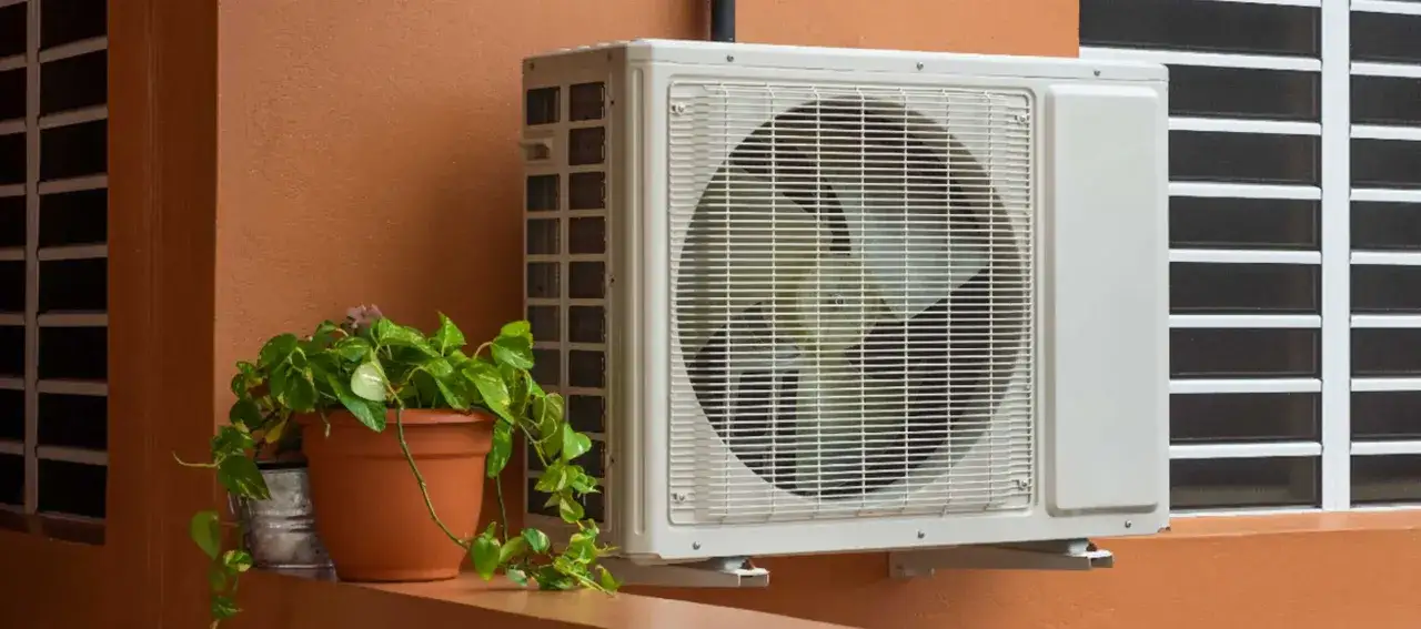 Unité extérieure de climatisation sur un mur orangé, à côté d'une plante. Faut-il éteindre sa pompe à chaleur l'été ?