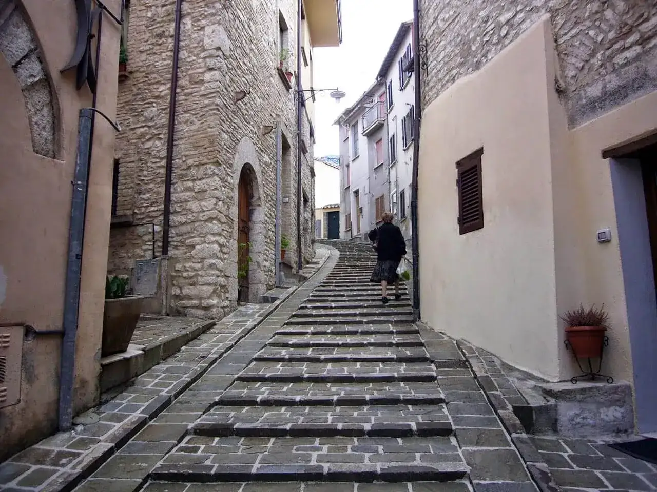 Una donna sale una scalinata in pietra in un vicolo di Gualdo Tadino, tra antiche case in pietra e intonaco chiaro.
