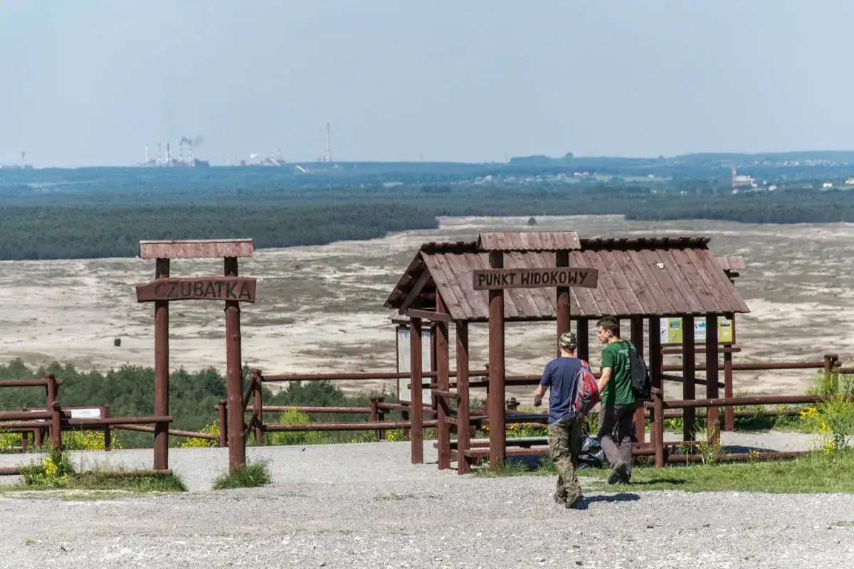 Dwaj turyści idą w stronę punktu widokowego na pustyni błędowskiej. W oddali widać przemysłowy krajobraz.