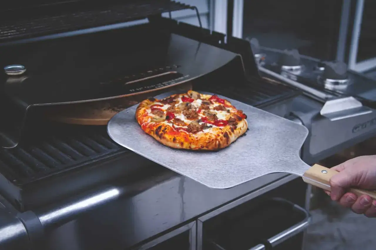Frisch gebackene Pizza wird mit einem Pizzaschieber aus einem Pizzaofen auf einem Gasgrill genommen.