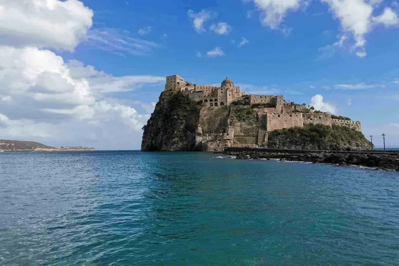 Castello Aragonese su uno scoglio, collegato alla terraferma da un ponte del 25 aprile. Acqua turchese e cielo azzurro con nuvole bianche.