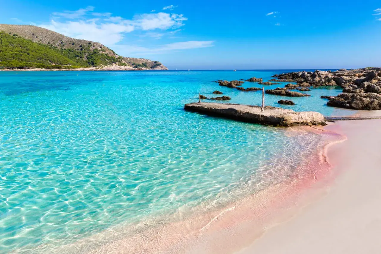Aguas cristalinas y arena rosada en una de las playas paradis&iacute;acas de Espa&ntilde;a. Un muelle de piedra se adentra en el mar turquesa.