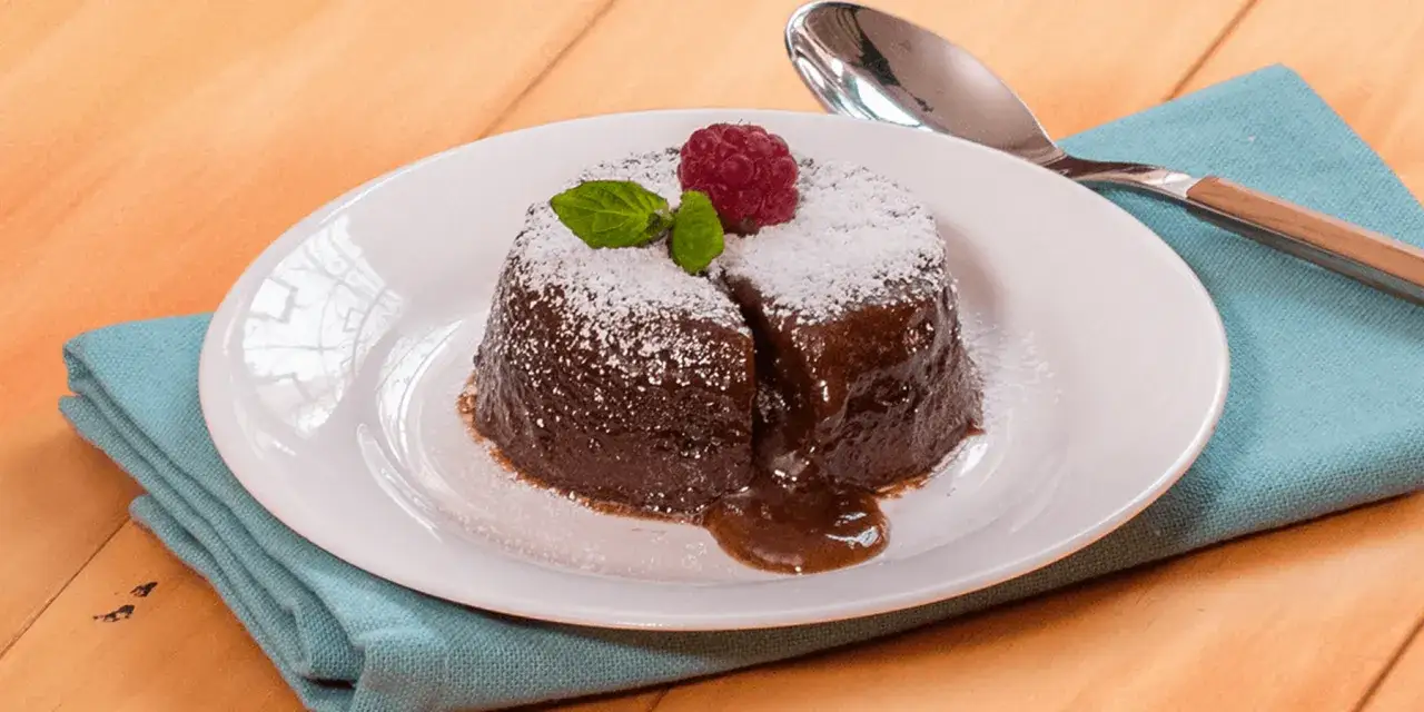 Coulant de chocolate reci&eacute;n horneado, cubierto de az&uacute;car glas, con una frambuesa y menta encima. El interior l&iacute;quido se desborda.