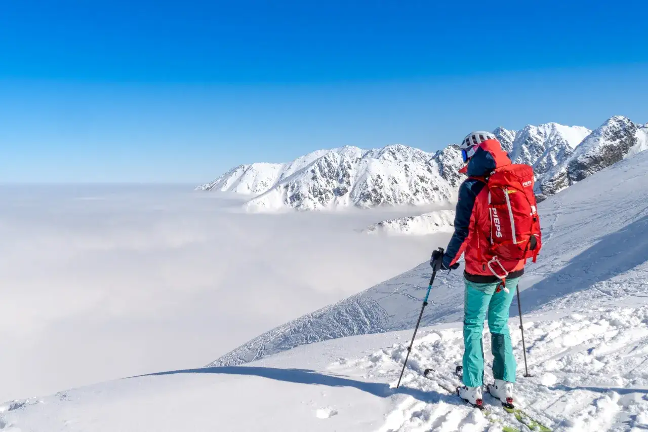 Skitury co to? Widok na ośnieżone góry nad morzem chmur. Narciarz w czerwonej kurtce i turkusowych spodniach z plecakiem PIPS podziwia panoramę.