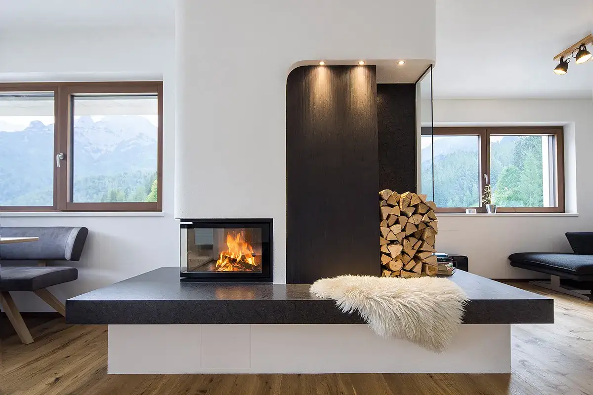 Moderner gemauerter Kamin mit brennendem Feuer, daneben ein Stapel Holz und eine Schaffell-Decke. Gro&szlig;e Fenster mit Bergblick.