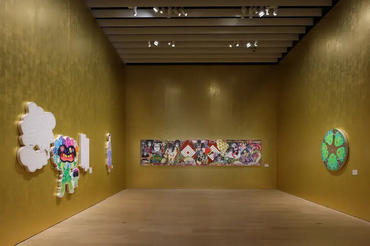 Takeshi Murakami's bunte Kunstwerke, darunter eine Wolke und ein Blumenwesen, h&auml;ngen an einer goldenen Wand neben einer langen, detailreichen Malerei und einem runden Baum-Bild.