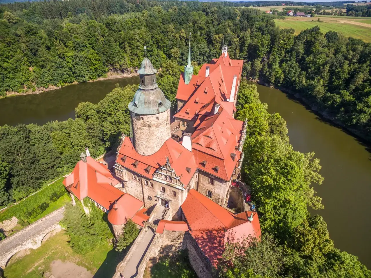 Zamek Czocha, otoczony zielenią i wodą, z czerwonymi dachami i wieżami, wygląda jak z bajki.