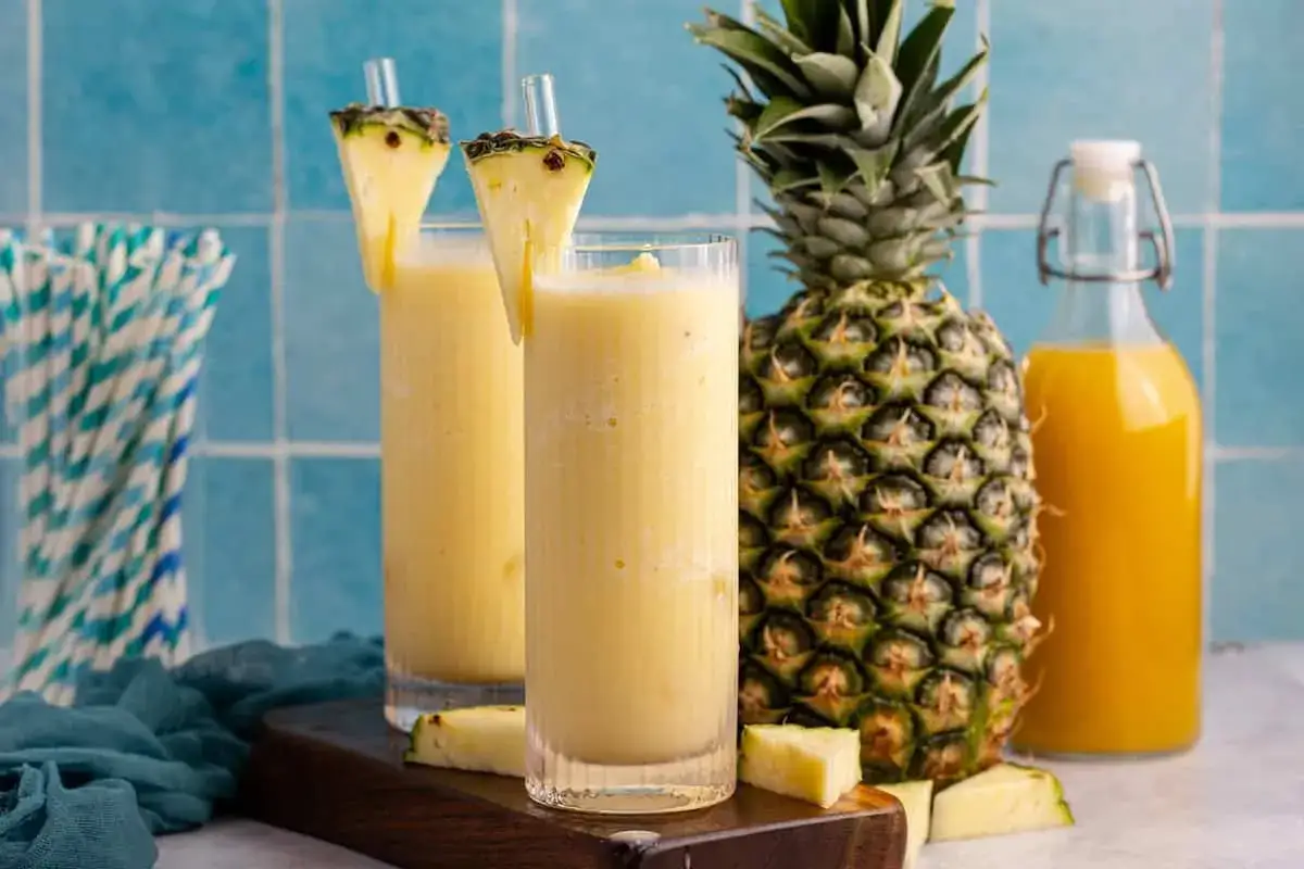 Zwei erfrischende Gläser alkoholfreier Piña Colada, garniert mit Ananasstücken, neben einer frischen Ananas und einer Flasche Saft.