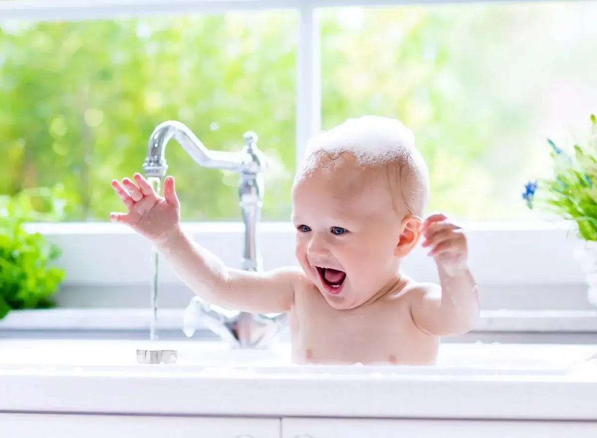 Ein fr&ouml;hliches Baby mit Schaum auf dem Kopf genie&szlig;t das Haare waschen in der Sp&uuml;le.