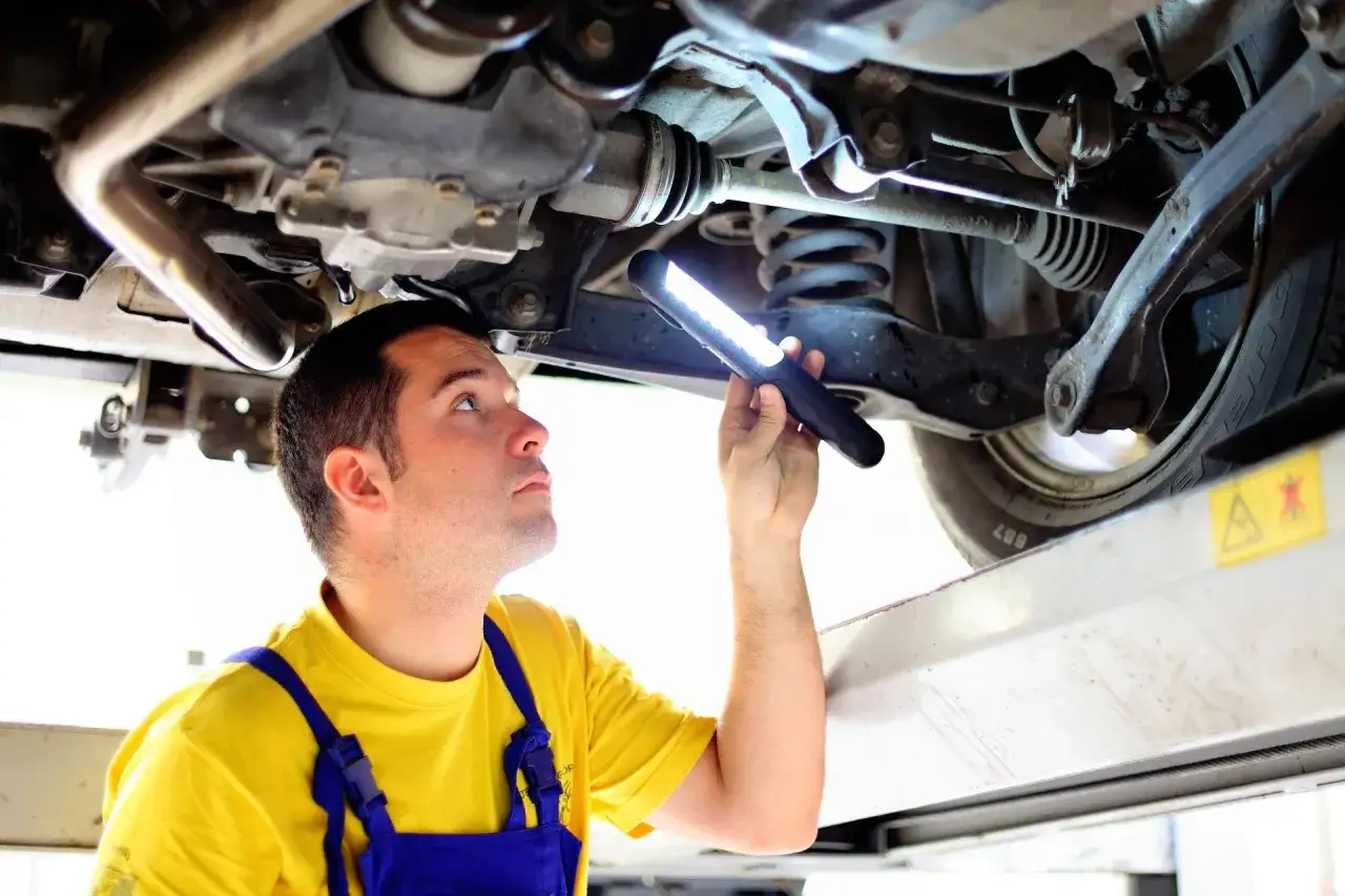 Mechanik szuka przyczyny metalicznego dźwięku podczas jazdy, oświetlając podwozie samochodu latarką.