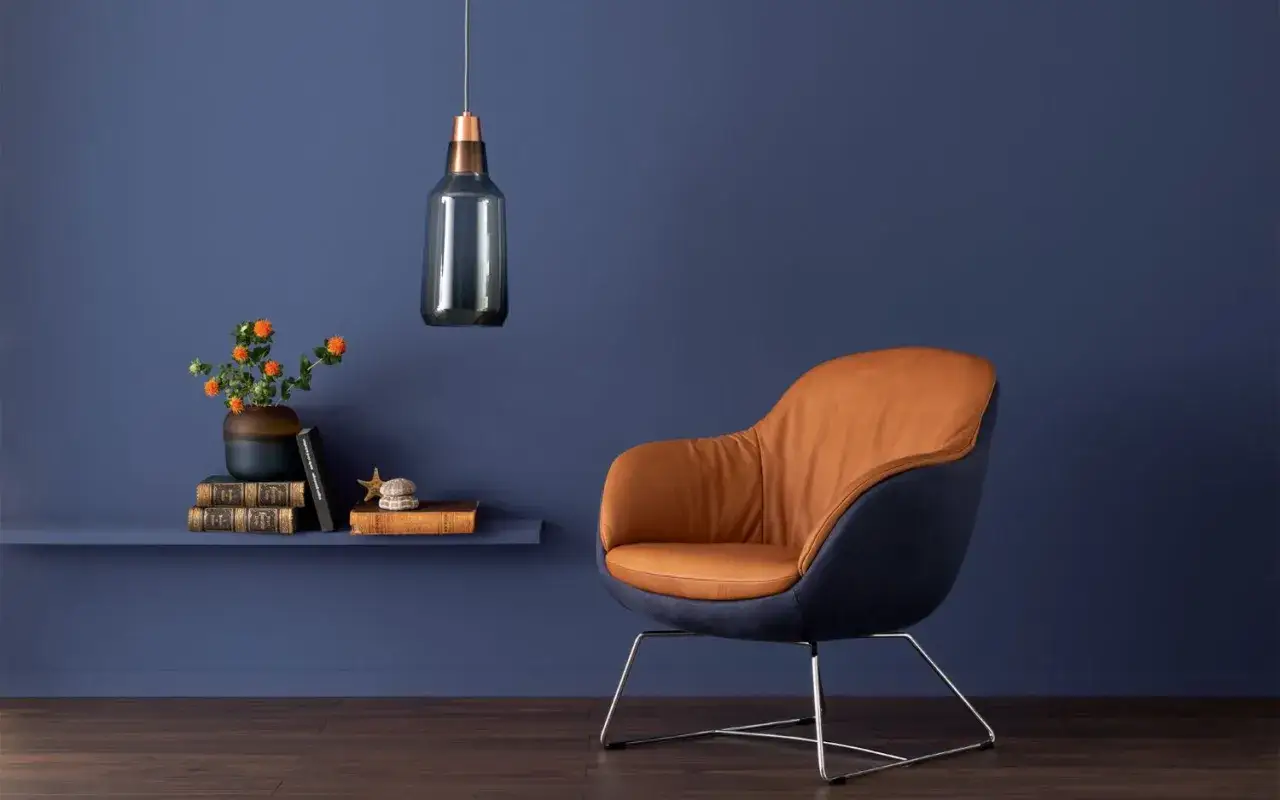 Ein Sessel mit cognacfarbenem Leder und dunkelblauem Stoff vor einer Wand in einem modernen Blau. Ein Regal mit Büchern und Blumen.
