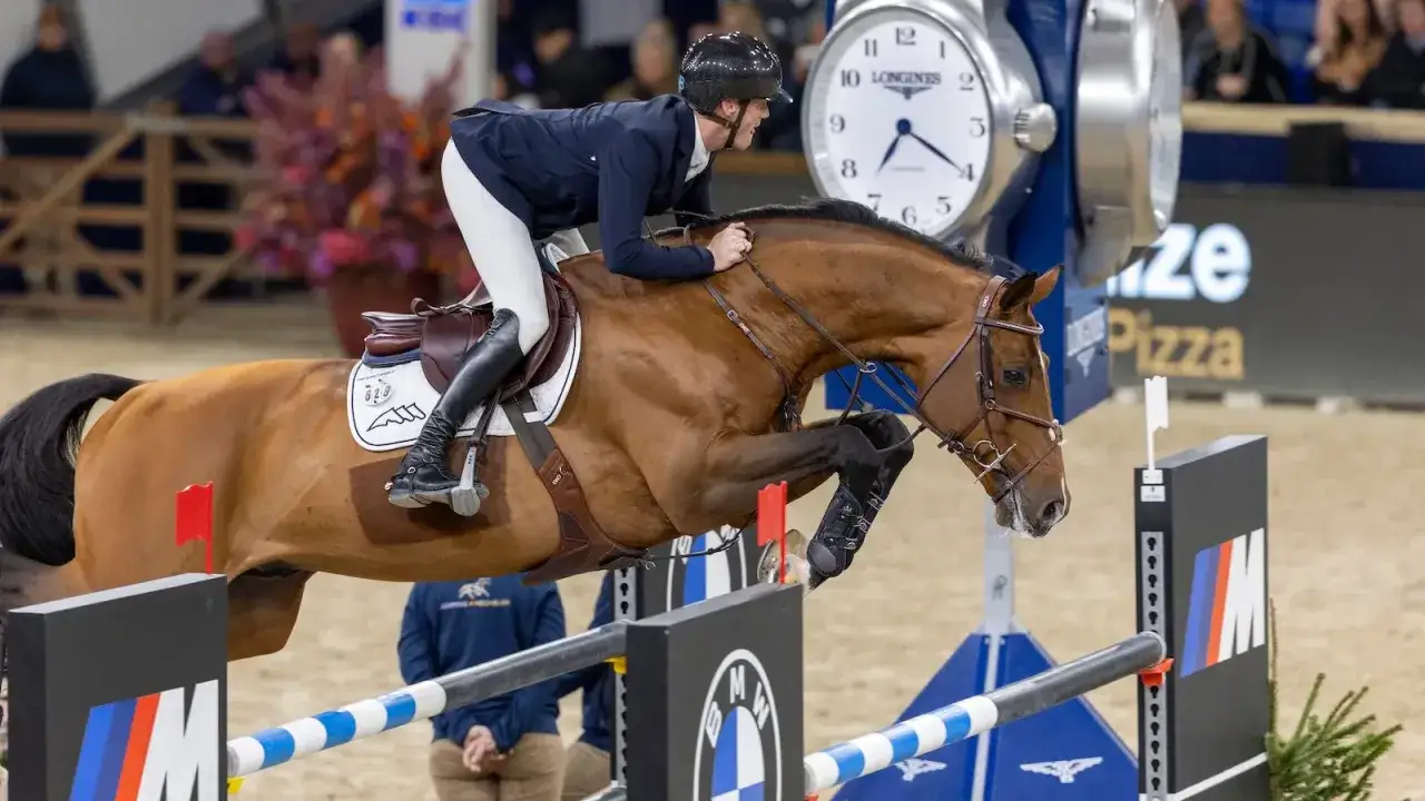 Un cavalier et son lando étalon brun sautent un obstacle. Une grande horloge Longines est visible en arrière-plan.