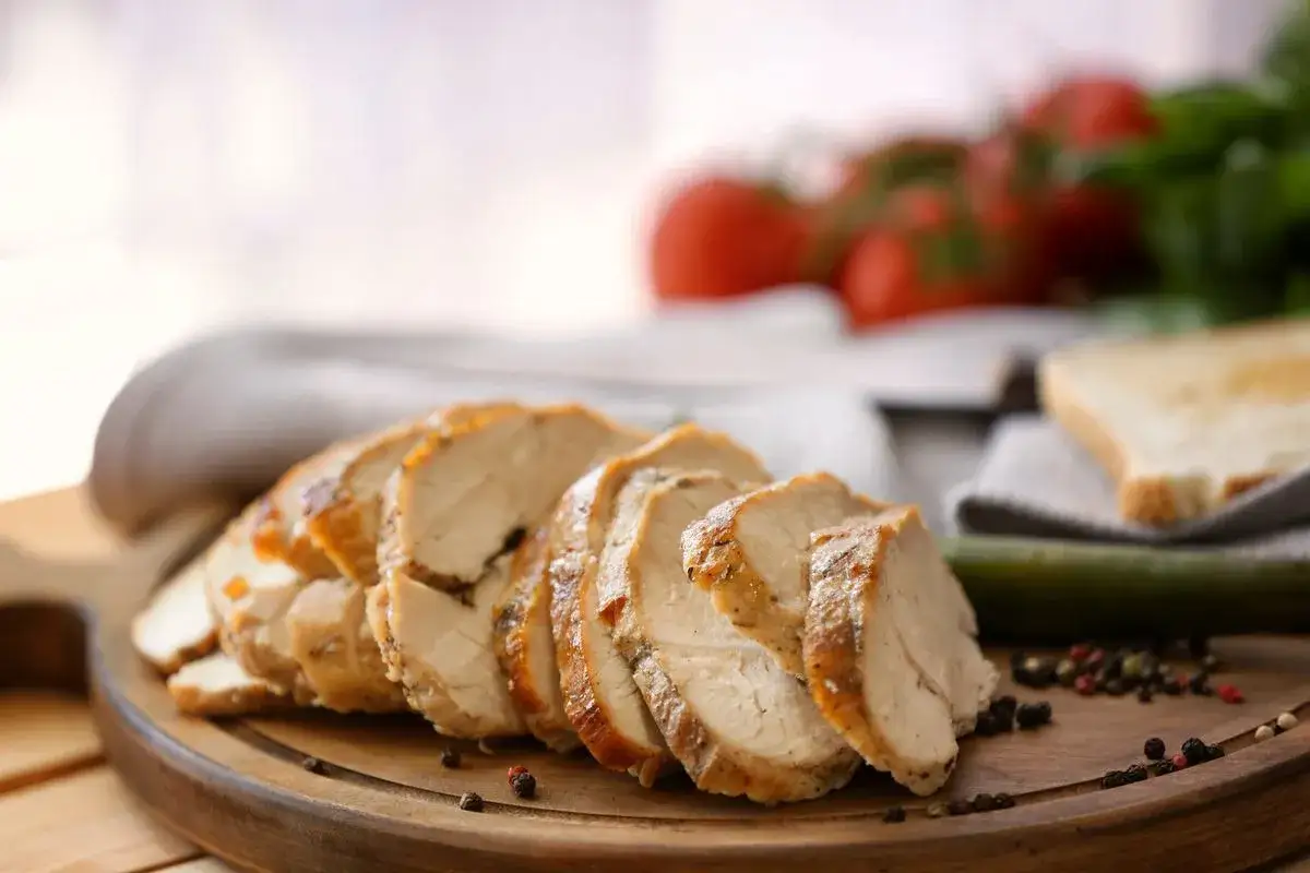 Fette di fesa di tacchino arrosto, condite con pepe, su un tagliere di legno, con pomodori e pane tostato sullo sfondo.