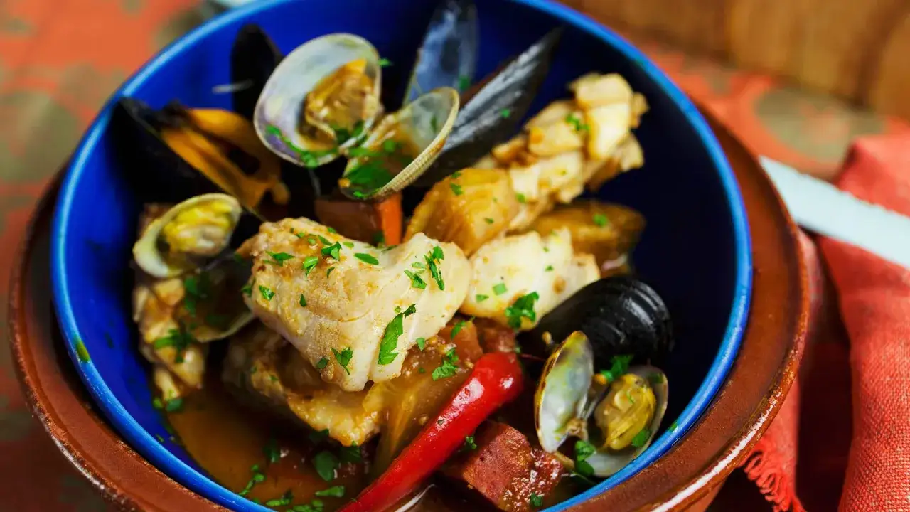 Un delicioso bacalao con almejas y gambas, adornado con perejil fresco y un toque de pimiento rojo.