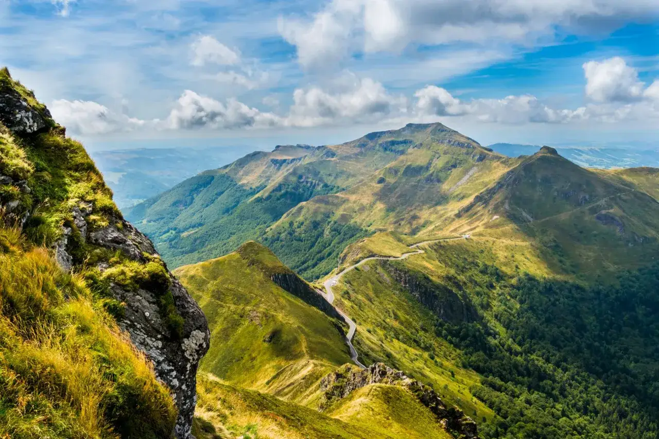 Vue panoramique des monts du Cantal, avec une route sinueuse traversant des paysages verdoyants sous un ciel nuageux.