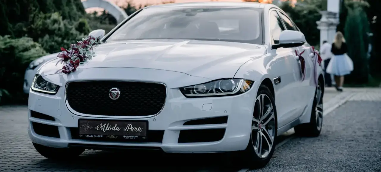 Elegancki biały Jaguar, ozdobiony na maskę i drzwi kwiatami, gotowy na przyjazd młodej pary.