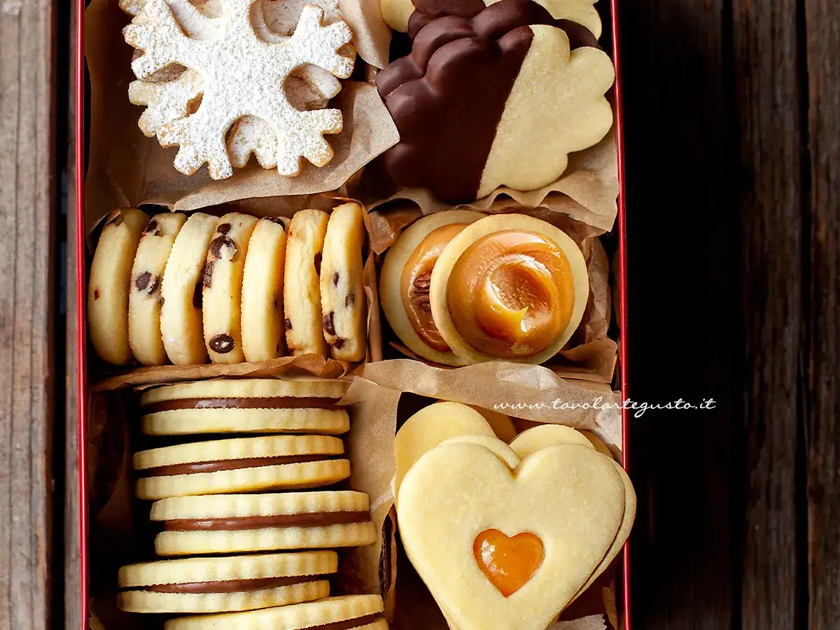 Scatola piena di biscotti al burro: a fiocco di neve, con gocce di cioccolato, ripieni di caramello, a forma di cuore e ricoperti di cioccolato.