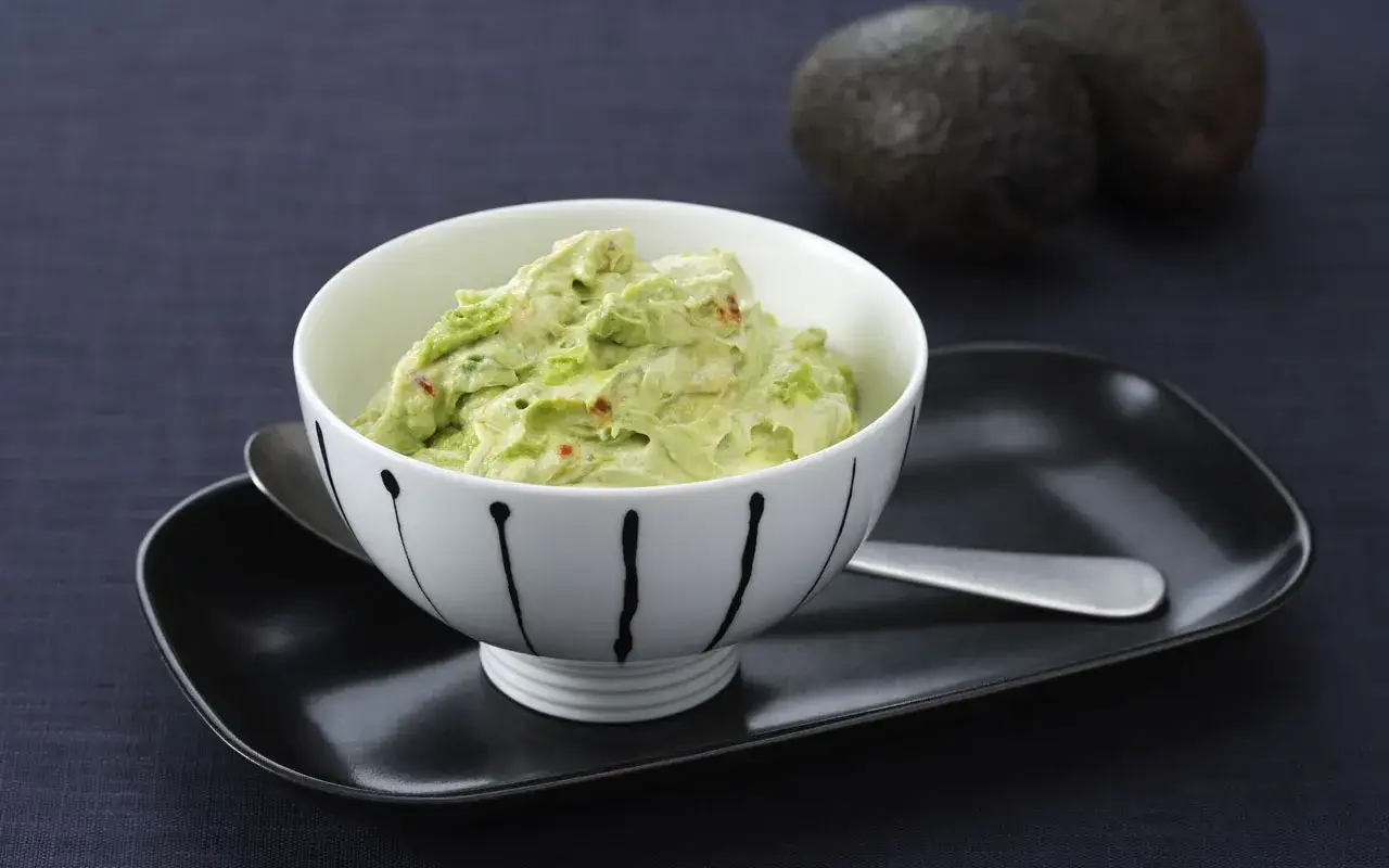 Kr&auml;mig guacamole med en touch av cr&egrave;me fra&icirc;che, serverad i en vacker sk&aring;l. Tv&aring; avokados syns i bakgrunden.