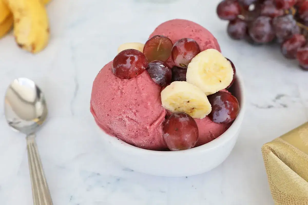 Schüssel mit cremigem Trauben Eis, garniert mit frischen Trauben und Bananenscheiben. Ein Löffel liegt daneben.