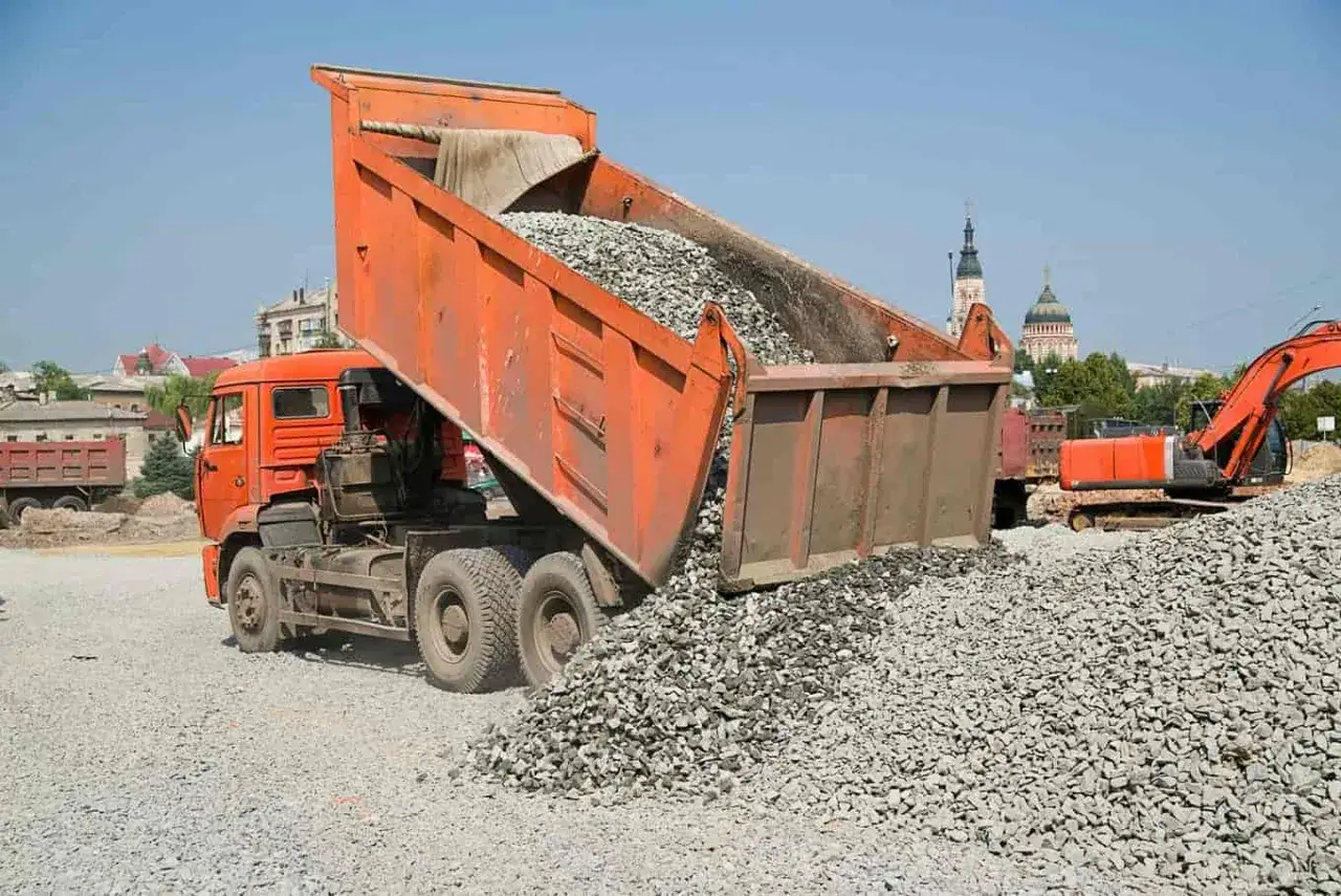 Pomarańczowa wywrotka wysypuje żwir. Zastanawiasz się, ile kosztuje tona żwiru? Na placu budowy widać koparkę i budynki.