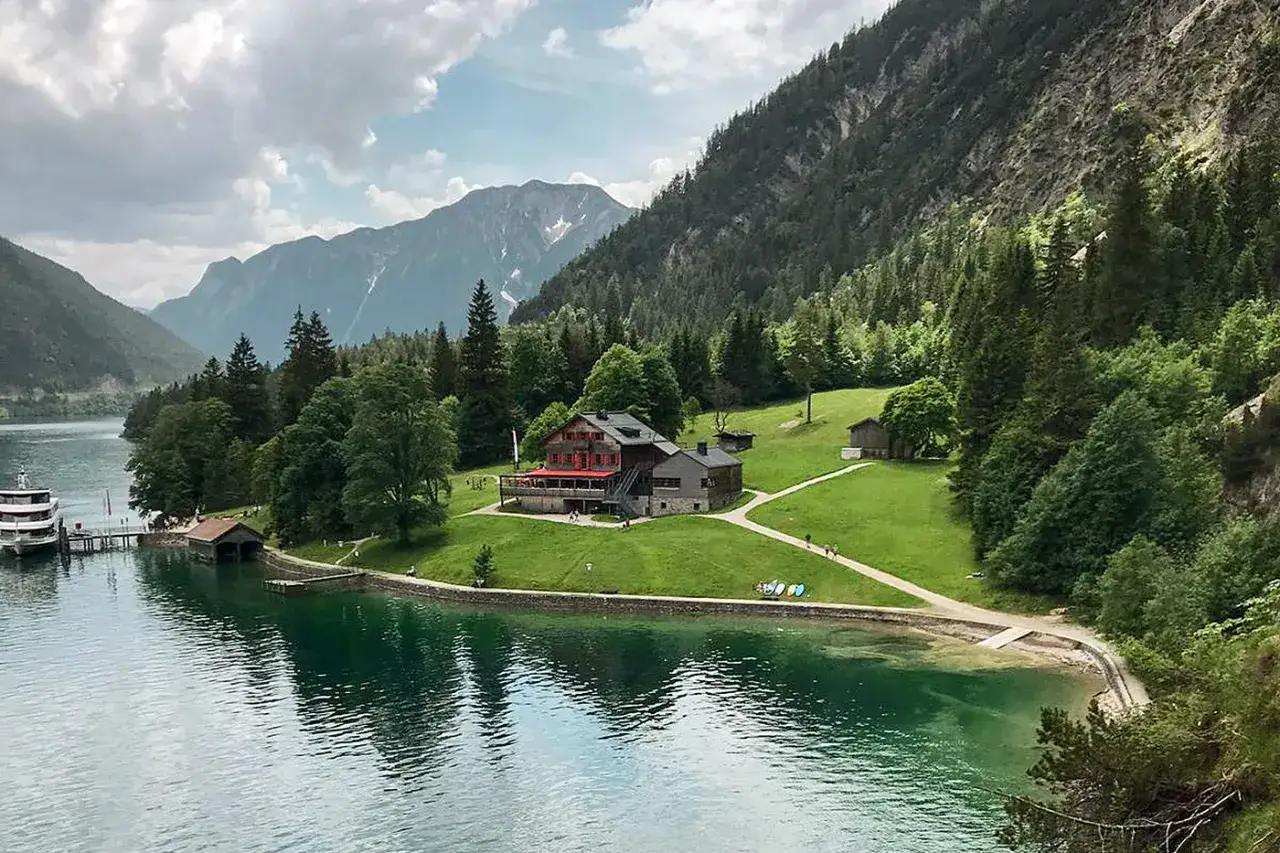 Gaisalm am Achensee: Ein Gasthaus mit roten Fensterl&auml;den liegt auf einer gr&uuml;nen Wiese am Ufer des t&uuml;rkisfarbenen Sees, umgeben von Bergen und W&auml;ldern.