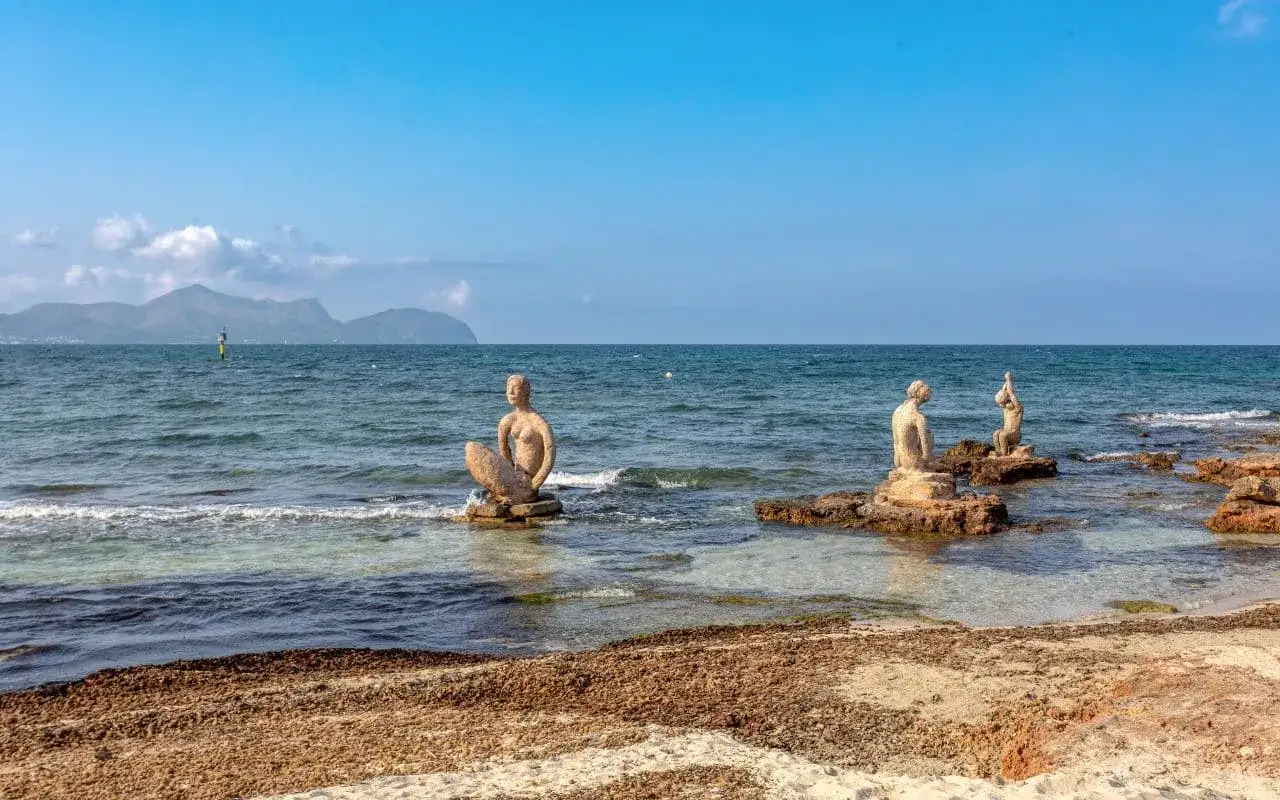 Skulpturen von Meerjungfrauen am Can Picafort Strand, Mallorca. Das Meer ist ruhig, im Hintergrund Berge.