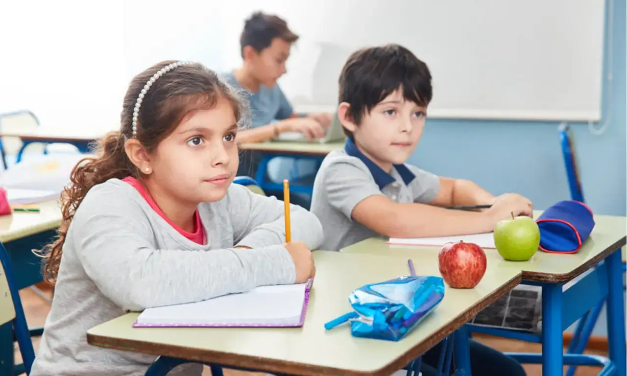 Zwei Kinder im Unterricht, bereit für einen Partnerdiktat. Ein Mädchen mit Perlenstirnband und ein Junge mit einem Apfel auf dem Tisch.