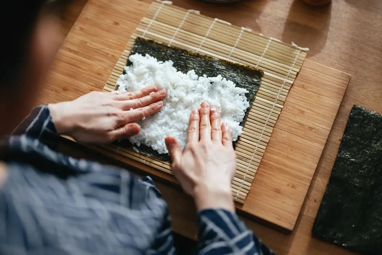 Ręce rozprowadzają ryż na nori, przygotowując się do zwinięcia sushi. Z 500g ryżu wyjdzie ok. 5-7 rolek.