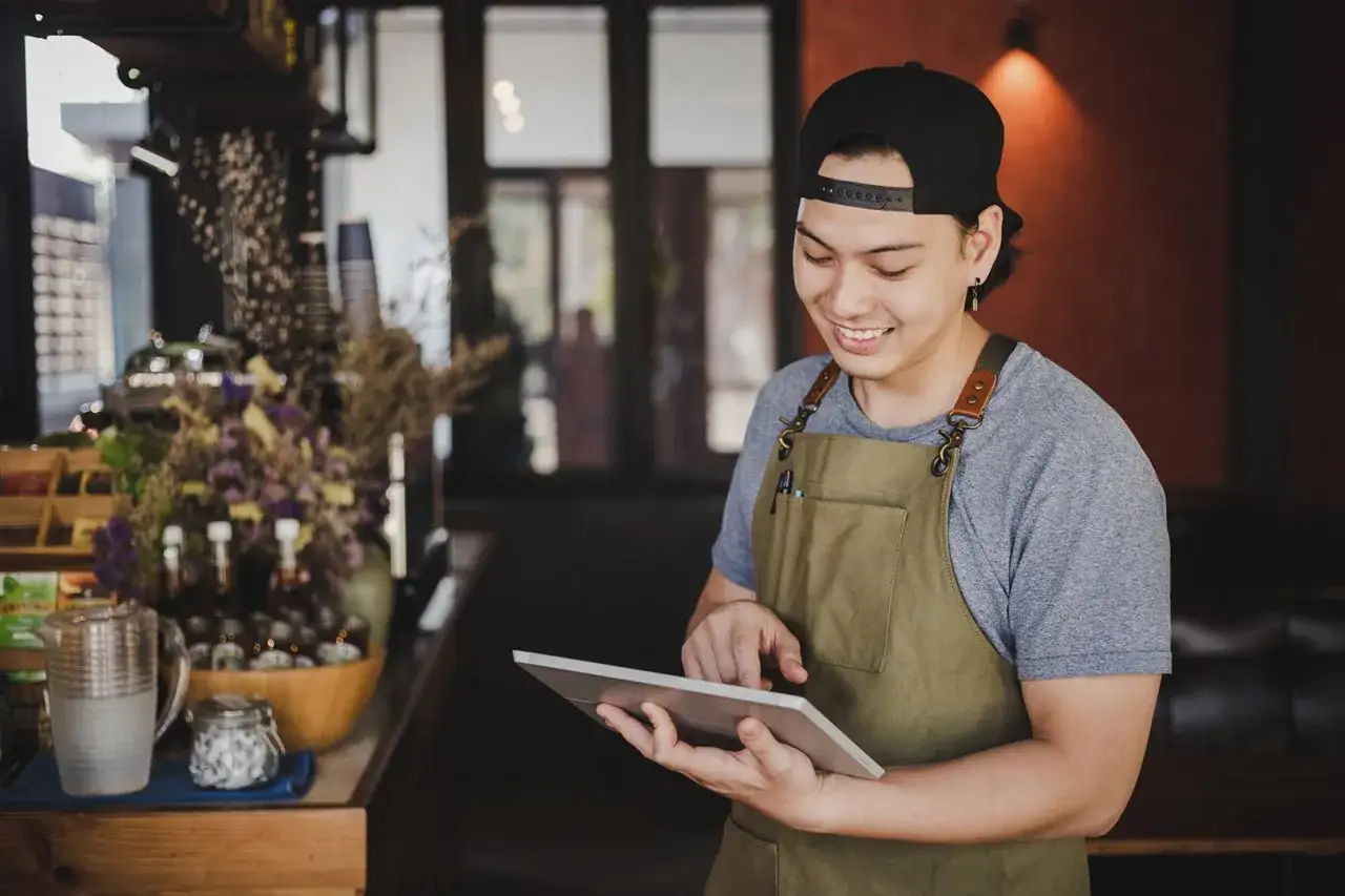 Lächelnder junger Mann mit Schürze und Kappe nutzt ein Tablet, ideal für einen werkstudenten job in einem Café.