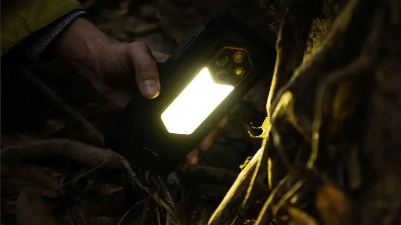 Ręka trzyma telefon survivalowy z włączoną mocną latarką, oświetlając leśne otoczenie.