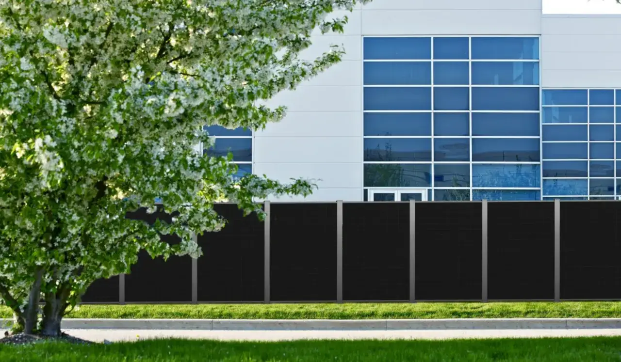 Ein moderner, schwarzer Zaun mit integrierten Solarmodulen vor einem Bürogebäude. Ein blühender Baum im Vordergrund. Ein solarzaun sinnvoll für Energie.