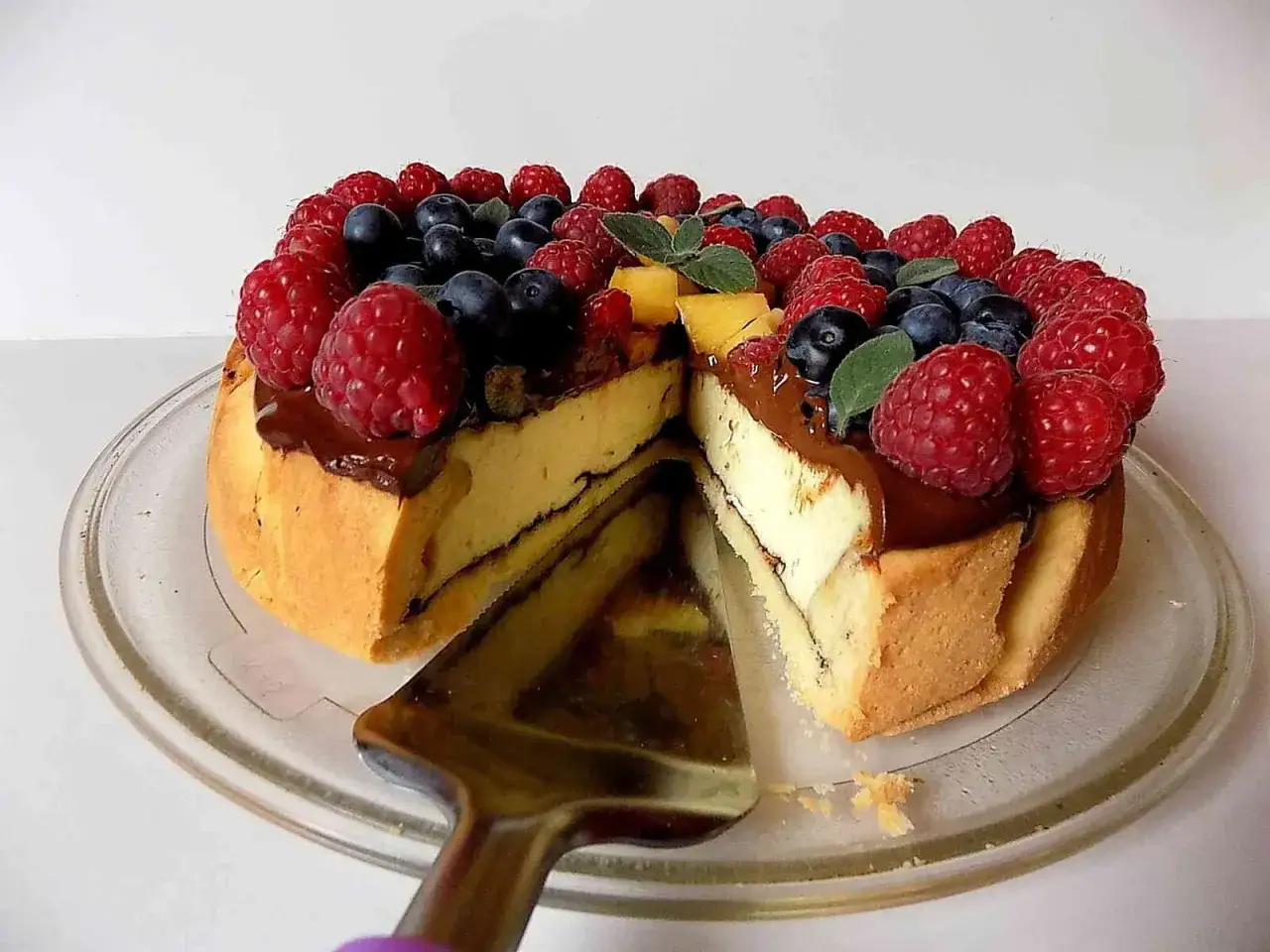 Sernik z polewą czekoladową, ozdobiony malinami, borówkami i kawałkami mango. Wygląda pysznie!