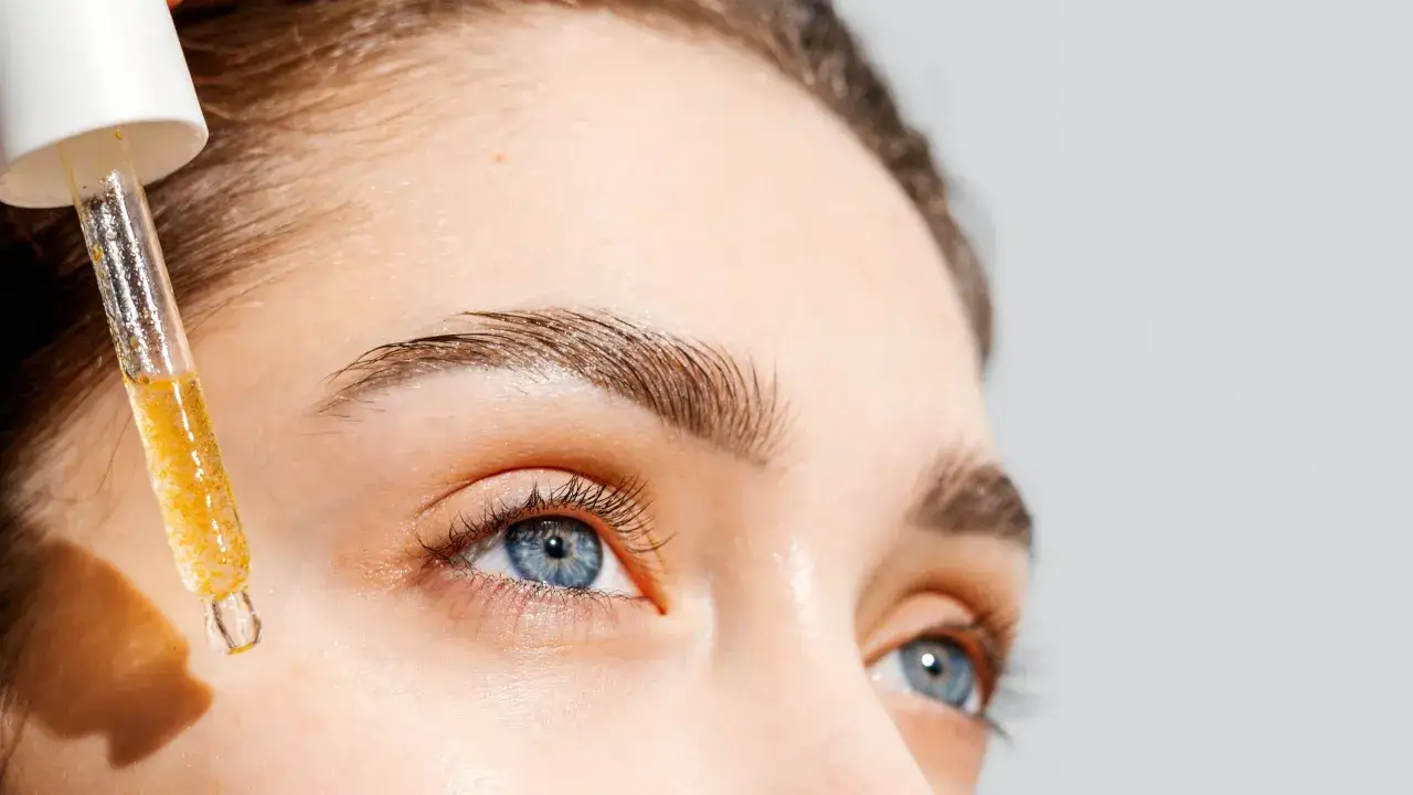 Ein Tropfen goldenes Vitamin C Serum für empfindliche Haut fällt von einer Pipette auf die Haut neben dem Auge.