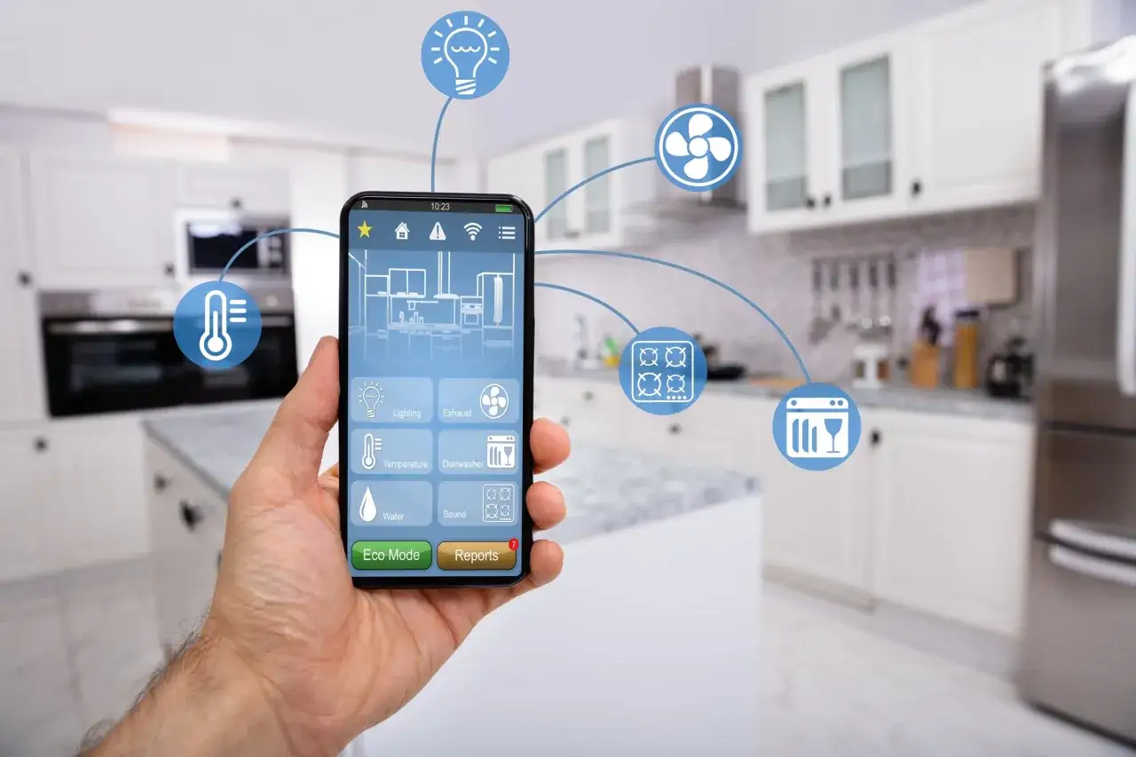 Dłoń trzyma smartfon z aplikacją do sterowania inteligentnymi urządzeniami w kuchni: światłem, wentylatorem, piekarnikiem, zmywarką i lodówką.