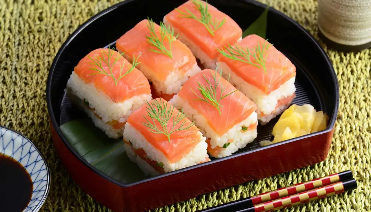Sześć kawałków sushi z ryżem i łososiem wędzonym na zimno, ozdobione gałązkami koperku. Obok imbir i sos sojowy.
