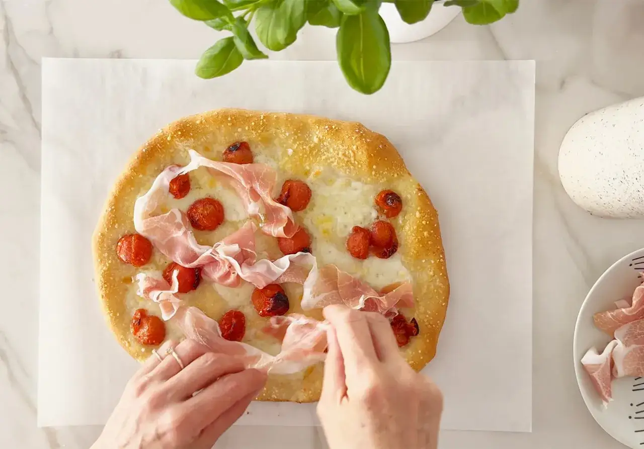 Hände legen dünne Scheiben **prosciutto schinken** auf eine Focaccia mit Kirschtomaten und Käse.