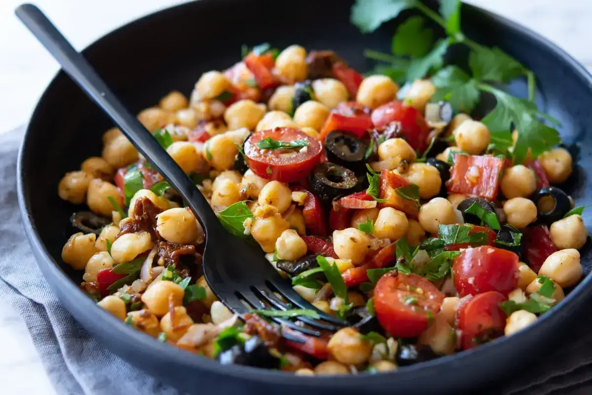 Un'insalata di ceci alla siciliana ricca di pomodorini, olive nere e prezzemolo fresco, servita in una ciotola scura con una forchetta.