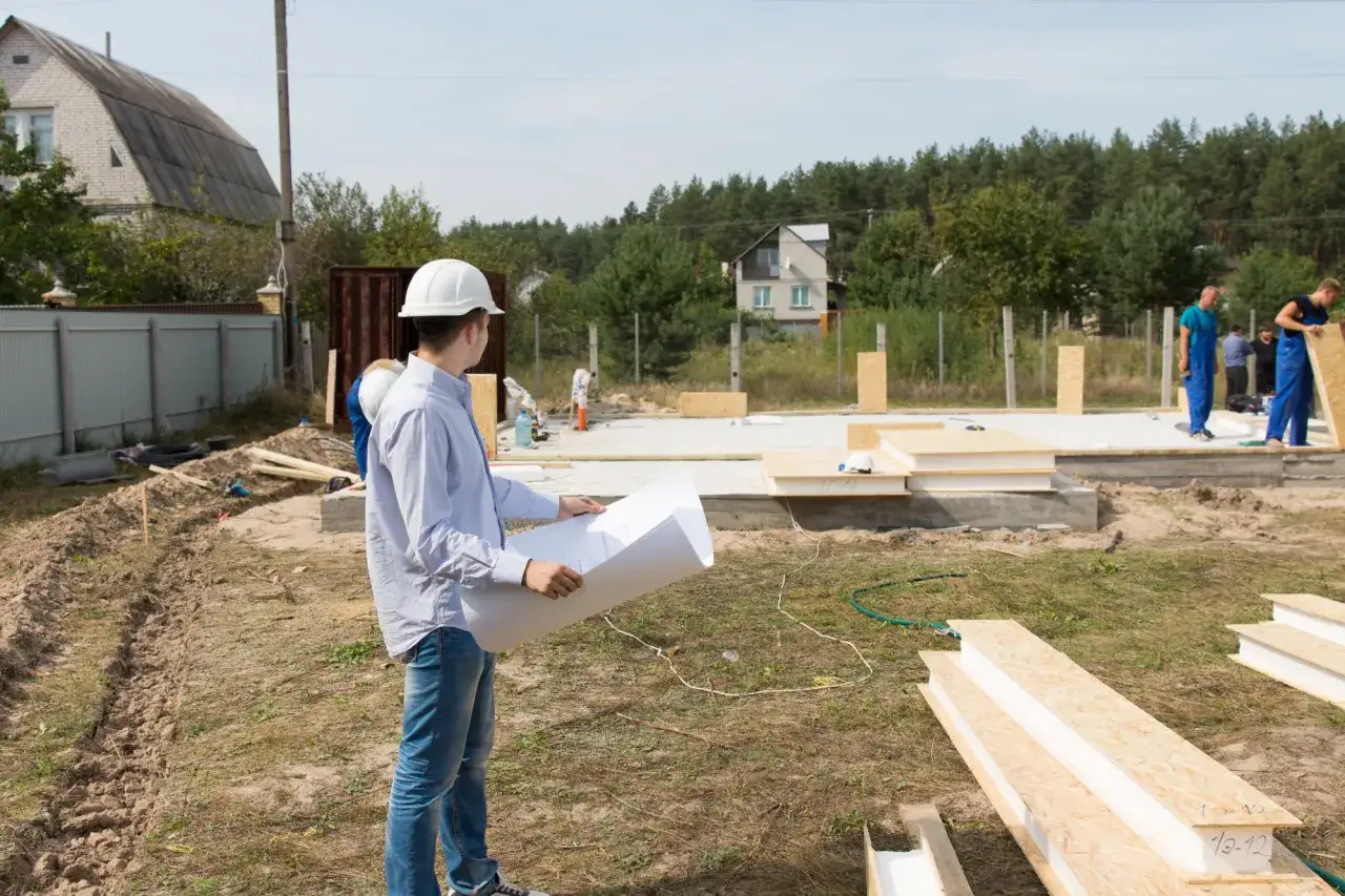 Budowa domu na wsi. Inżynier analizuje plany, sprawdzając minimalną wielkość działki budowlanej. Pracownicy montują elementy konstrukcyjne.