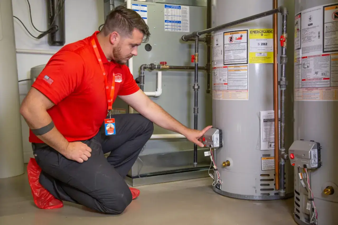Technician in red shirt and boot covers inspects a water heater, possibly to replace it.
