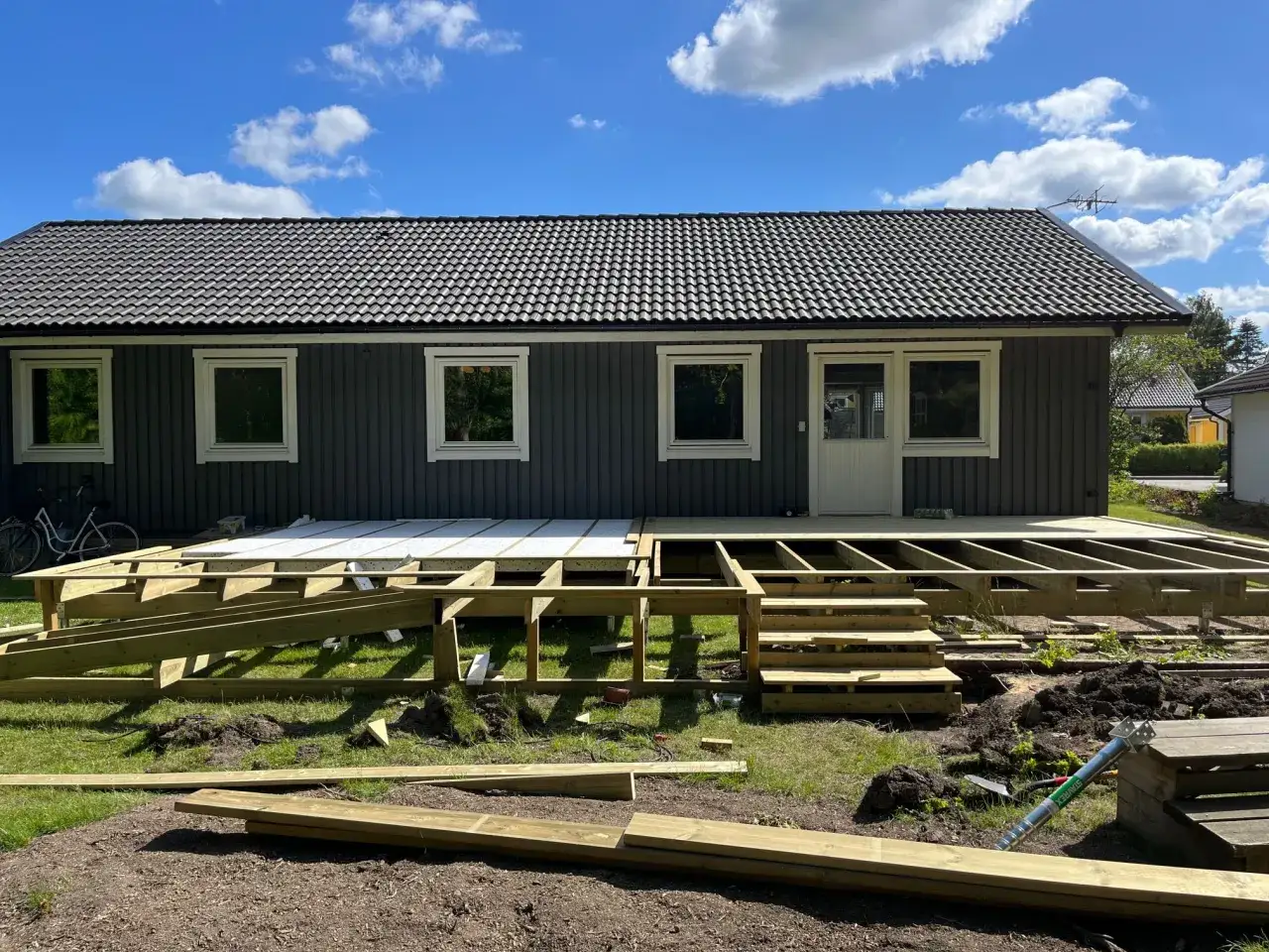 Bygga altan: Träkonstruktion för en ny altan tar form framför ett mörkt trähus. Solen skiner på en blå himmel med moln.