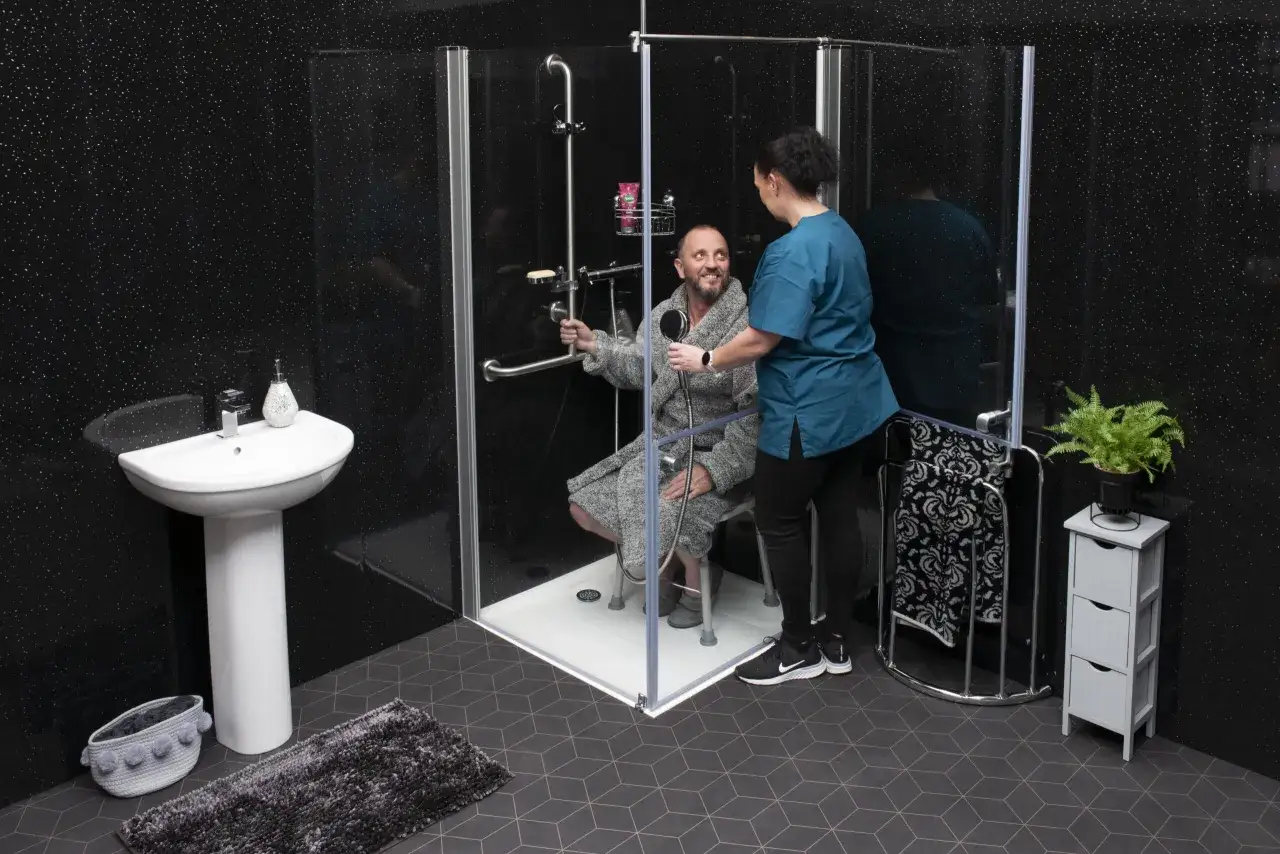 Une aide-soignante aide un homme âgé dans une douche adaptée. La salle de bain est équipée de barres d'appui et d'un siège pour faciliter l'aide salle de bain senior.
