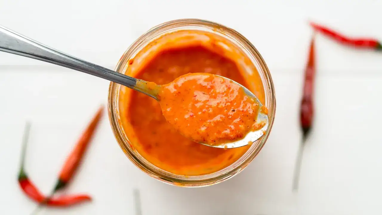 En sked med kryddig piri piri-sås lyfts ur en burk. Runt burken syns röda chilifrukter.