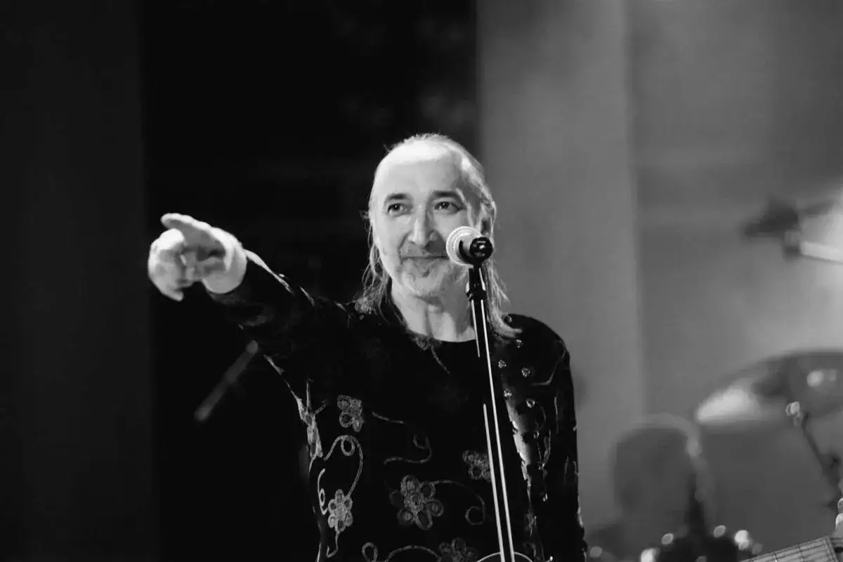 Artysta w czarnej, wzorzystej koszuli wskazuje palcem, śpiewając do mikrofonu. W tle widać instrumenty, oprócz błękitnego nieba.