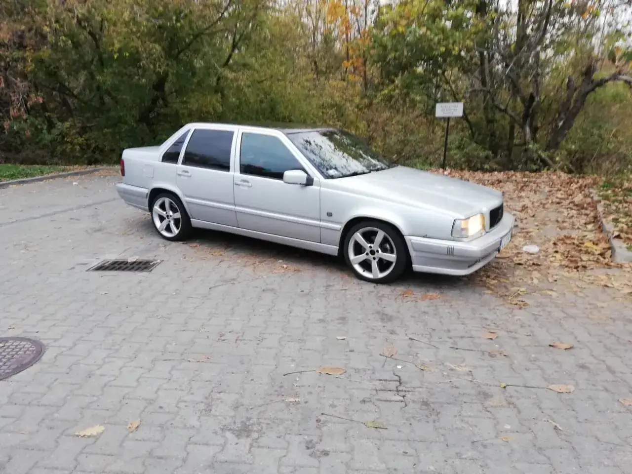 Srebrne Volvo 840 z przyciemnianymi szybami stoi na brukowanym placu, otoczone jesiennymi liśćmi i drzewami.