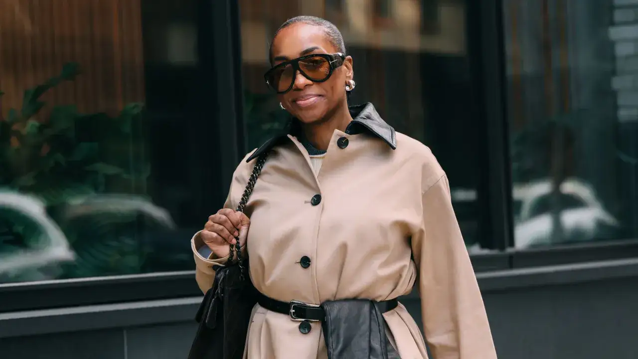 A woman in a tan trench coat with a black leather collar and belt, sporting large sunglasses, embodies chic 2026 fashion trends.