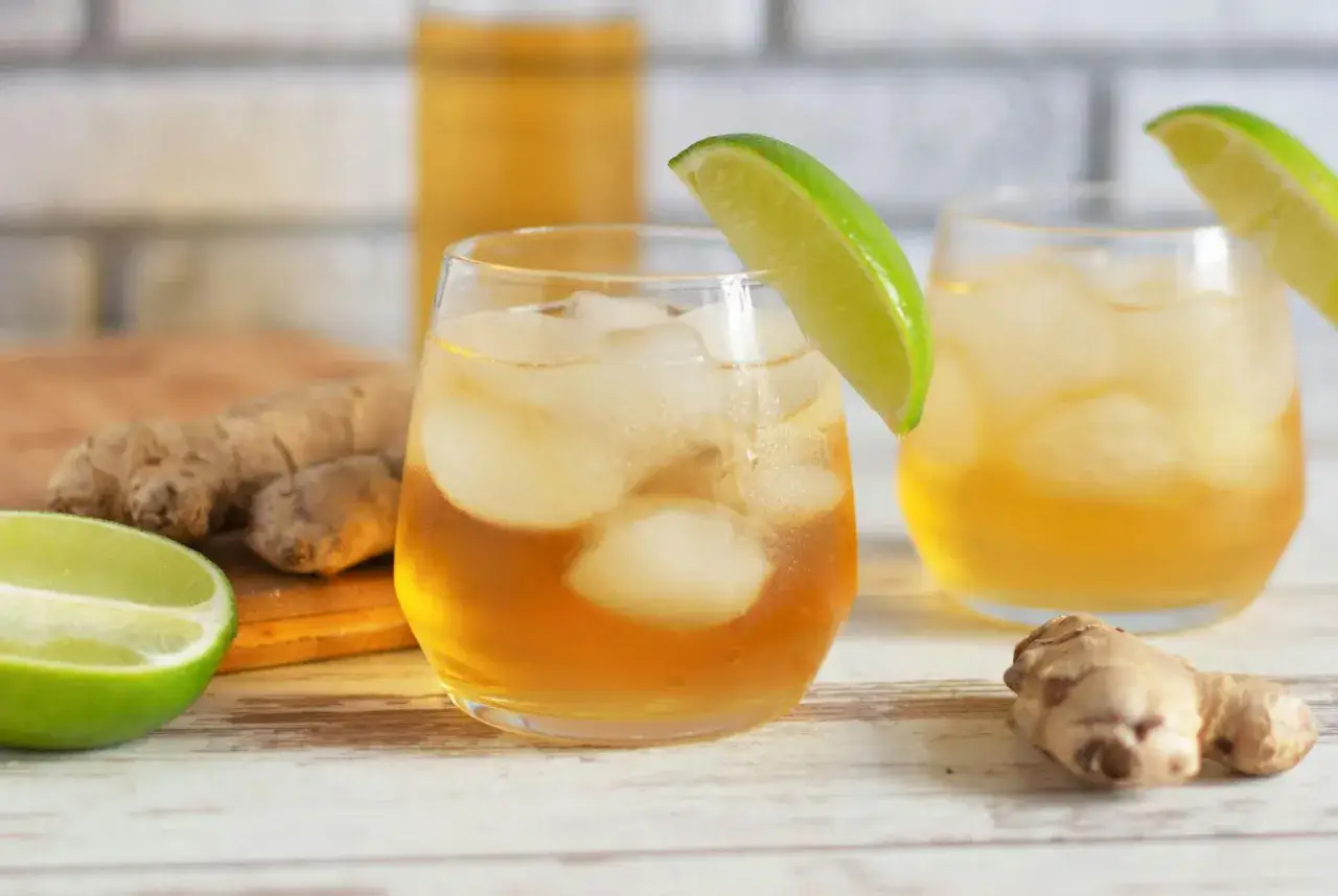 Två glas med iskyld ginger beer, garnerade med limeklyfta. Färska ingefärskvistar och lime i bakgrunden.