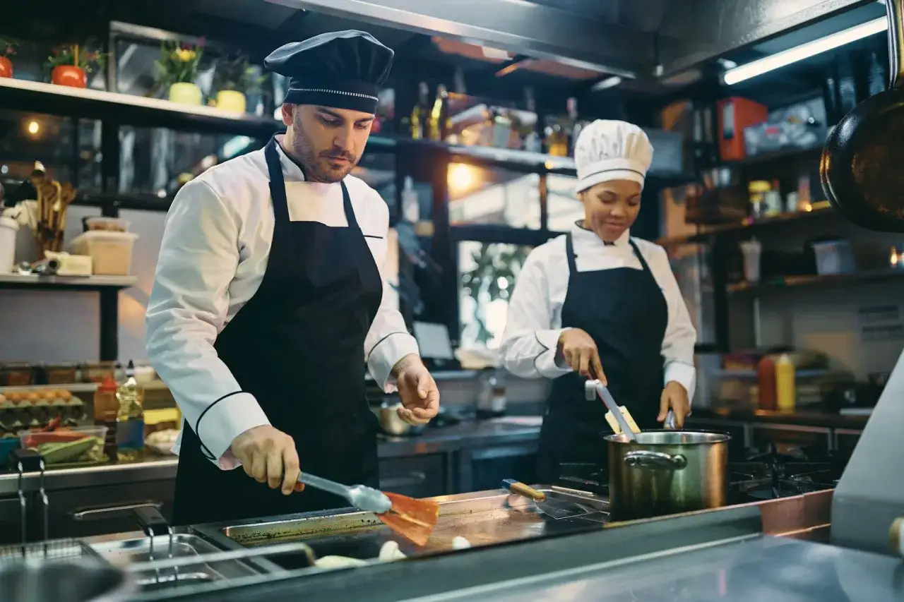 Dwóch kucharzy w kuchni restauracyjnej przygotowuje posiłek. Mężczyzna z przodu używa łopatki, kobieta z tyłu miesza coś w garnku. Kod zawodu kucharz.