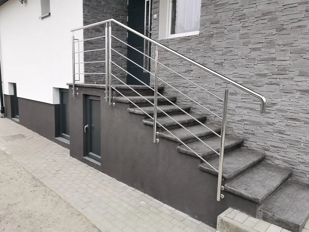 Nowoczesna, **jaka balustrada na schody zewnętrzne** ze stali nierdzewnej, idealnie komponuje się z kamienną elewacją i betonowymi stopniami.