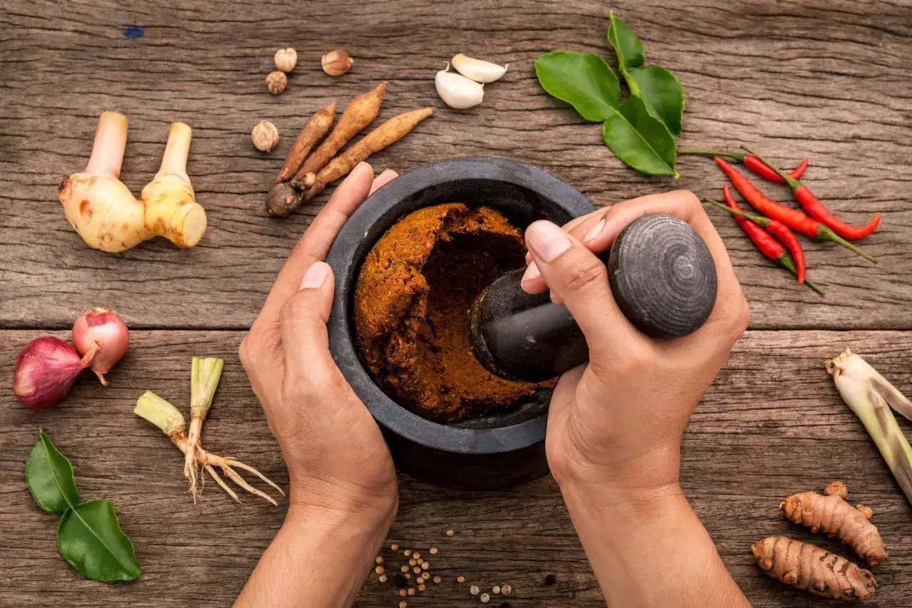 H&auml;nde zermahlen mit einem M&ouml;rser frische Zutaten f&uuml;r Currypaste. Ingwer, Chili, Knoblauch und Kr&auml;uter liegen bereit.